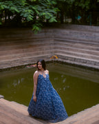 A woman wearing a blue floral print maxi dress stands on the stone steps next to a pond in an outdoor garden setting.
