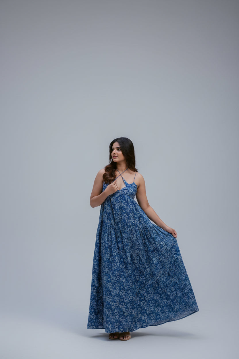 A woman wearing a blue floral print maxi dress stands by a stone pool in an outdoor park setting with trees and stone steps in the background.