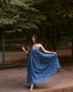A woman wearing a blue floral print maxi dress stands outdoors on a dirt path, holding out the sides of the dress to show its flow. The background features a green chain-link fence and dense green trees.